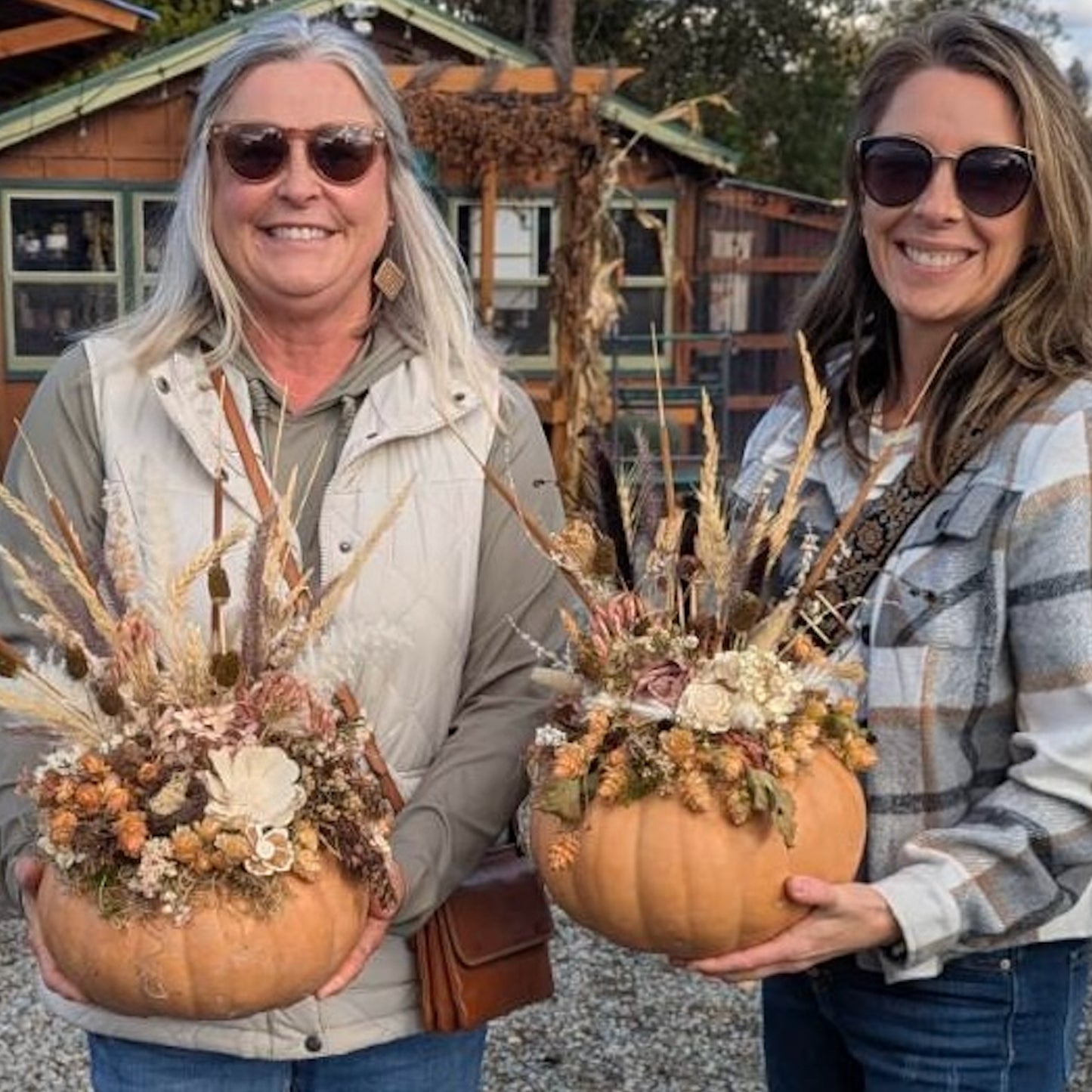 Two women holding decorative pumpkins with floral arrangements in front of a wooden cabin.