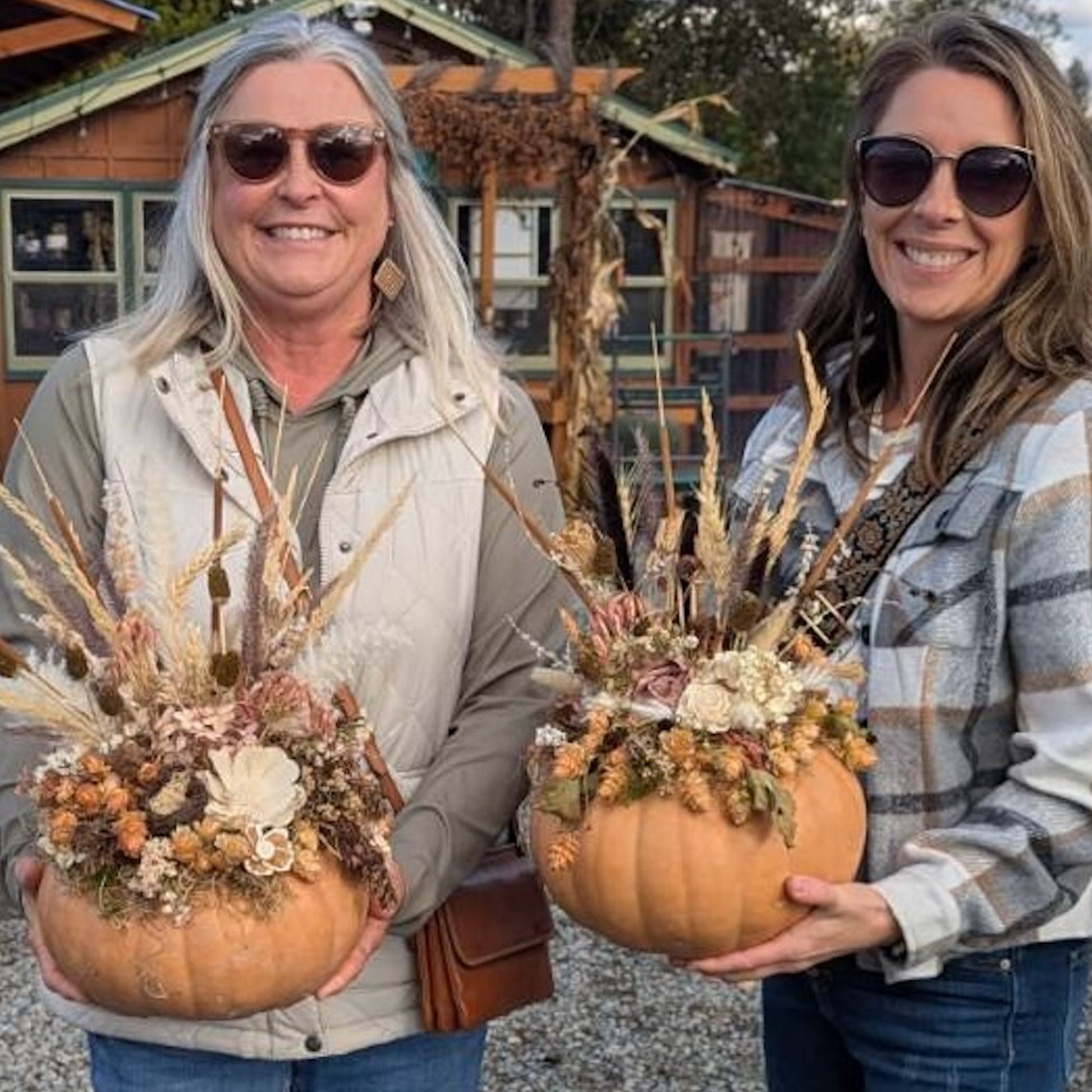 Two women holding decorative pumpkins with floral arrangements in front of a wooden cabin.